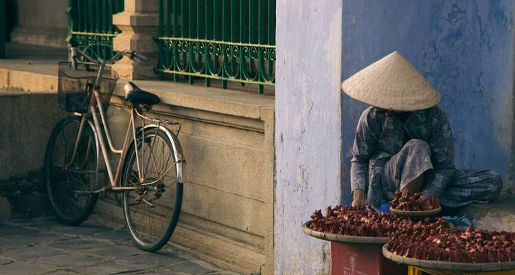 Verkoper met een kegelvormige hoed die goederen verkoopt bij een fiets, tegen een blauwe muur.