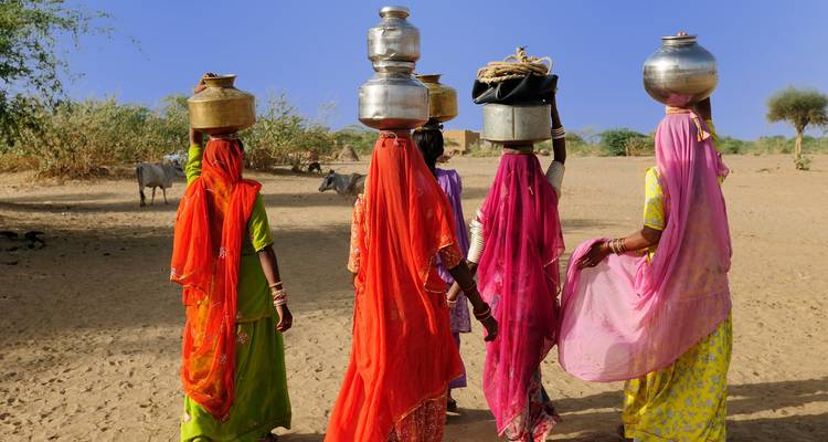 Gruppe von traditionell gekleideten Frauen, die Töpfe auf ihren Köpfen tragen, in einer Wüstenlandschaft.