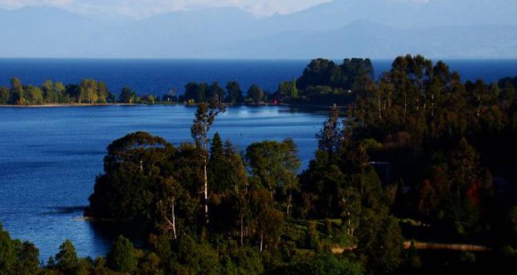 Panorama sereno a orillas del lago con penínsulas boscosas y montañas lejanas bajo cielos azules despejados.
