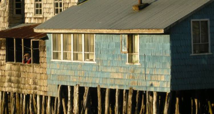 Casa de madera desgastada sobre pilotes con tejas azules, un viajero solitario fotografiando a través de una ventana abierta.