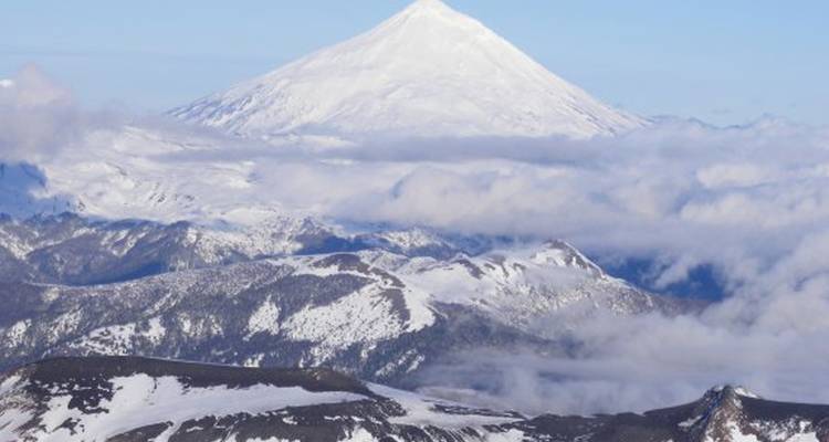Volcán cubierto de nieve rodeado de nubes y montañas.