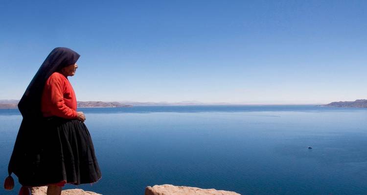Indigene Frau in traditioneller Kleidung steht auf einem felsigen Vorsprung und blickt auf die tiefblaue Weite eines Hochgebirgssees unter klarem Himmel.
