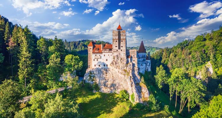 Castillo de Bran sobre una roca rodeado de árboles bajo un cielo azul.