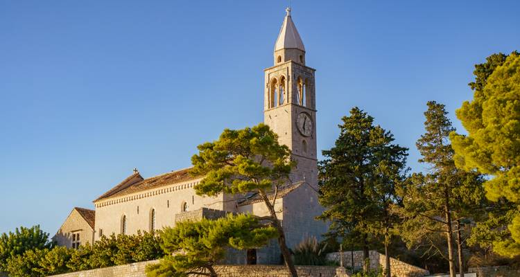 Église en pierre avec un haut clocher et une horloge baignés de lumière dorée chaude parmi des cyprès.