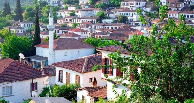 Hillside Turkish village of white houses with red-tiled roofs surrounded by green trees on a sunny day.