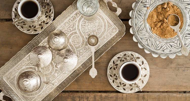 Overhead view of ornate silver tray set with Turkish coffee cups, water glass and brown sugar on rustic wood.