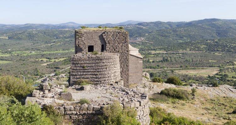 Stone tower ruins sit atop a rocky hill overlooking wide rural valleys and distant mountains under hazy light.