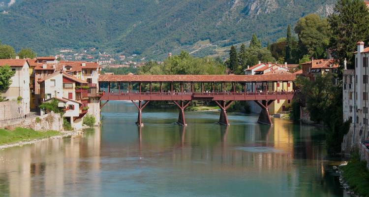 Historische overdekte houten brug die een groene rivier overspant met alpine heuvels op de achtergrond.