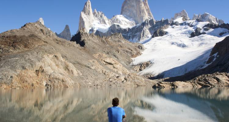 Individuo sentado junto a un lago reflectante con montañas de fondo.