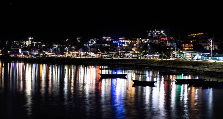 Paisaje urbano junto al lago nocturno con luces de colores reflejándose en el agua tranquila y botes silueteados