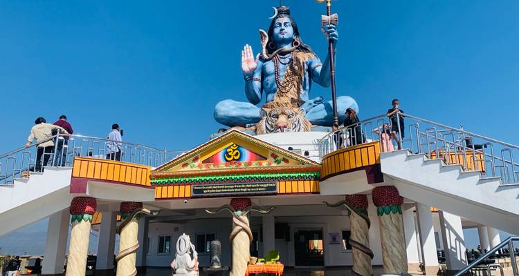 Gran estatua azul del Señor Shiva encima de una plataforma de templo con visitantes subiendo las escaleras bajo un cielo despejado