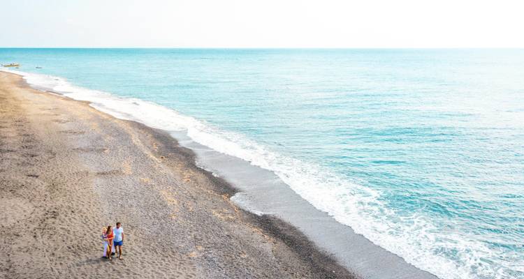 Pareja joven caminando de la mano por una amplia playa de guijarros vacía junto al mar turquesa