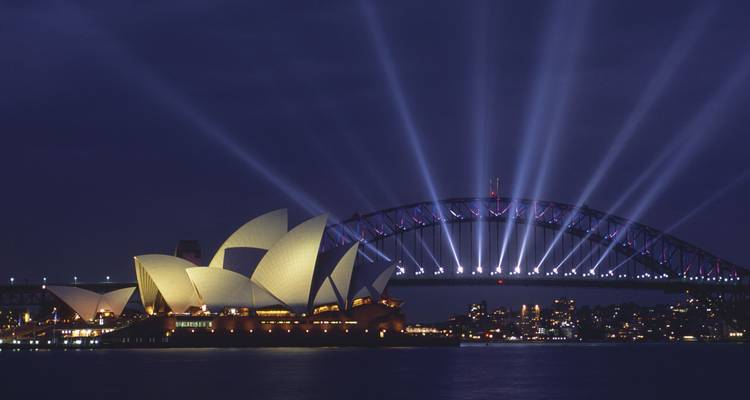 Sydney Opera House and Harbour Bridge illuminated by powerful night spotlights over the harbour.