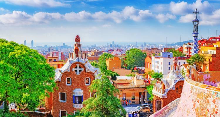 Bright morning view across Park Güell’s gingerbread gatehouses toward Barcelona cityscape and sea.