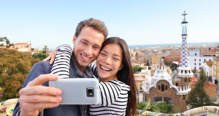 Smiling couple taking a selfie with Park Güell’s mosaic towers and Barcelona skyline behind them.