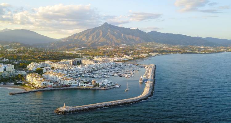 Aerial of luxury marina and curved breakwater with Marbella’s Sierra Blanca mountains beyond.
