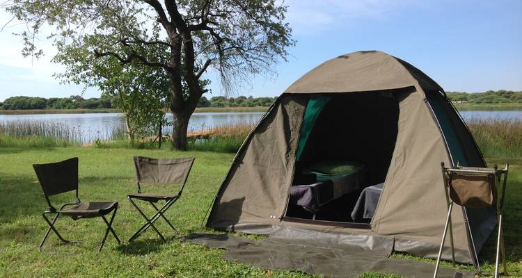 Tienda de campaña instalada junto a un lago con sillas de camping.