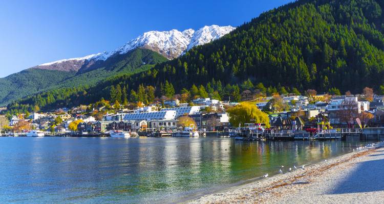 Crystal clear Lake Wakatipu reflecting snow-capped peaks and forested slopes beside Queenstown waterfront.