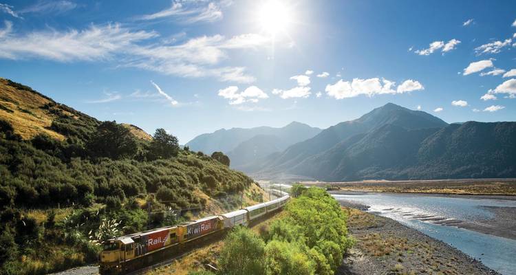 Long passenger train winds through a sunlit alpine valley beside a glacial river and towering peaks.