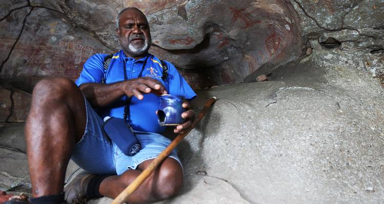 Un anciano aborigen demostrando artefactos dentro de una cueva adornada con arte rupestre antiguo.