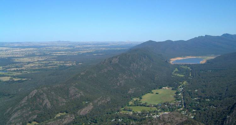 Vista panorámica de la cresta sobre llanuras amplias, laderas boscosas y un embalse distante bajo cielos despejados.