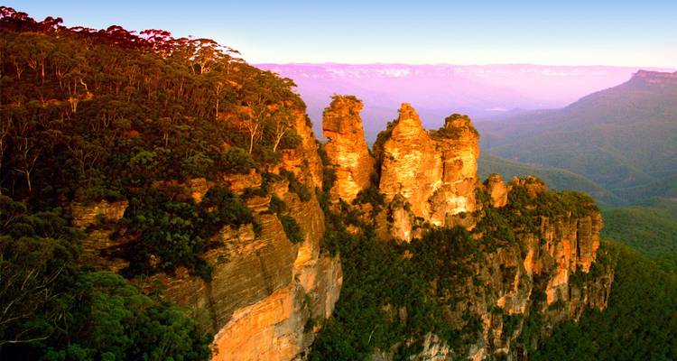 Los tres pilares de roca de arenisca Three Sisters brillando de color naranja al atardecer sobre un profundo valle verde.