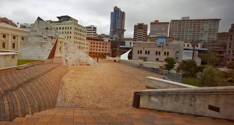 Plaza cívica moderna con sendero de ladrillos, esculturas de concreto angulares y edificios de la ciudad bajo cielos grises.