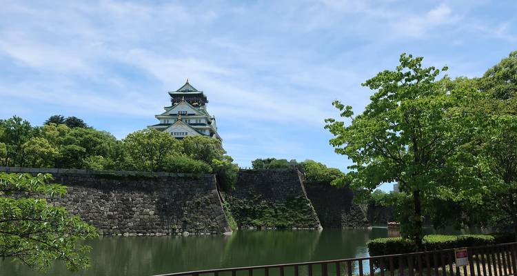 Le château d'Osaka s'élève au-dessus de murs en pierre et d'un fossé réfléchissant entouré d'un feuillage vert éclatant et d'un ciel bleu.