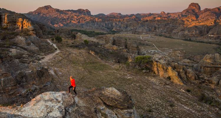 Un randonneur solitaire vêtu d'une chemise rouge se tient sur une corniche rocheuse surplombant le paysage spectaculaire du canyon avec ses formations de grès illuminées par une lumière dorée.