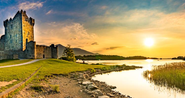 Coucher de soleil sur les ruines d'un château au bord d'un lac avec des montagnes en silhouette à l'arrière-plan.