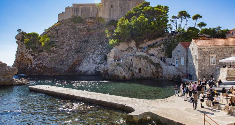 Les touristes se rassemblent sur un quai en pierre sous une forteresse surplombant une falaise qui domine des eaux côtières cristallines.