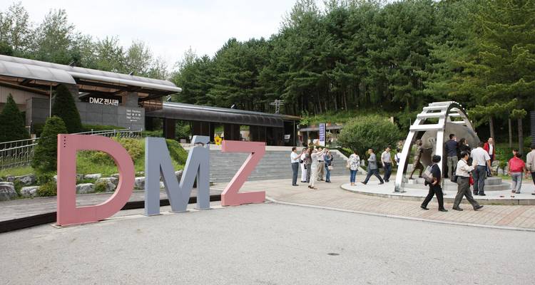 Entrada del área de tour de la DMZ con grandes letras de DMZ y turistas reunidos cerca del bosque de pinos.