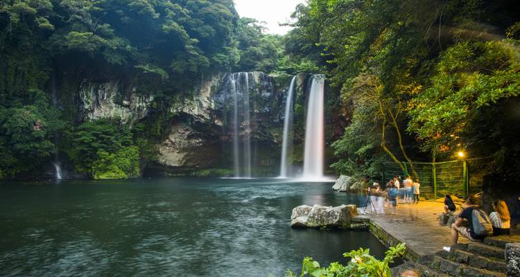 Tranquila cascada cayendo en una piscina de esmeraldas rodeada de un frondoso bosque con visitantes en la plataforma de observación al atardecer.