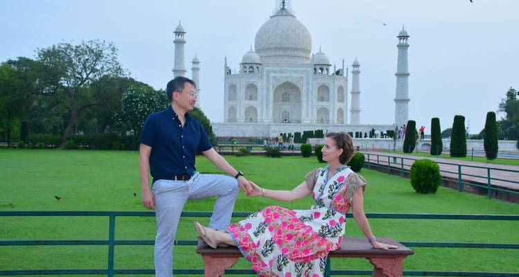Una pareja posando de forma juguetona en un banco con el Taj Mahal de fondo en un jardín verde