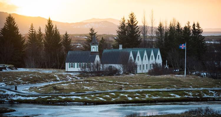 Petite église avec un drapeau islandais au coucher du soleil, entourée d'arbres et de neige.