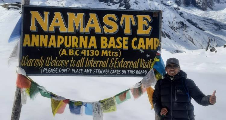 Person am Annapurna Basislager mit einem Willkommensschild und schneebedeckten Bergen.