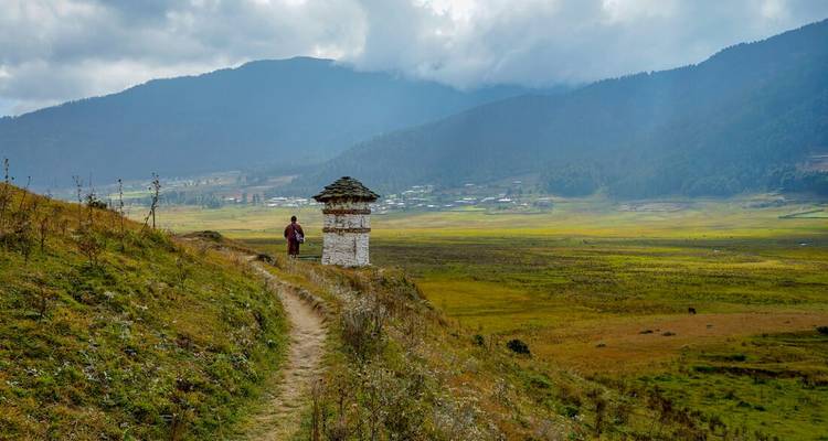 Persoon die langs een pad loopt met traditionele Bhutanese structuur en bergen op de achtergrond.