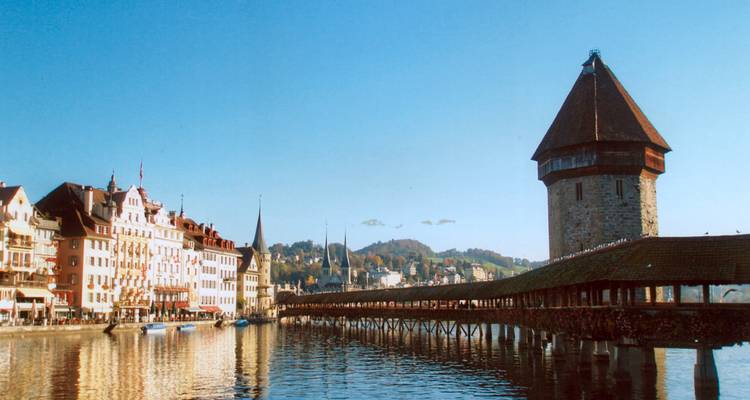 Le Pont de la Chapelle et sa tour en pierre se reflétant dans les eaux calmes de Lucerne avec des bâtiments fluviaux aux couleurs vives.