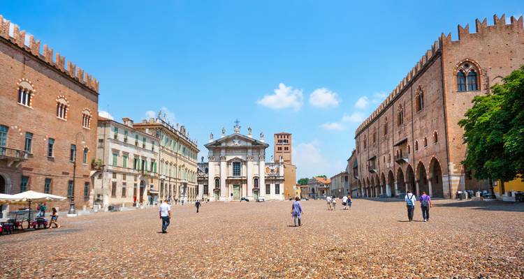 Large piazza pavée de pavés flanquée de palais historiques et d'une église à Mantoue avec quelques personnes se promenant sous un ciel bleu.