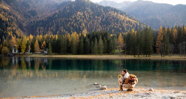 Young man crouching with his dog beside a turquoise alpine lake surrounded by autumn forest and mountains.