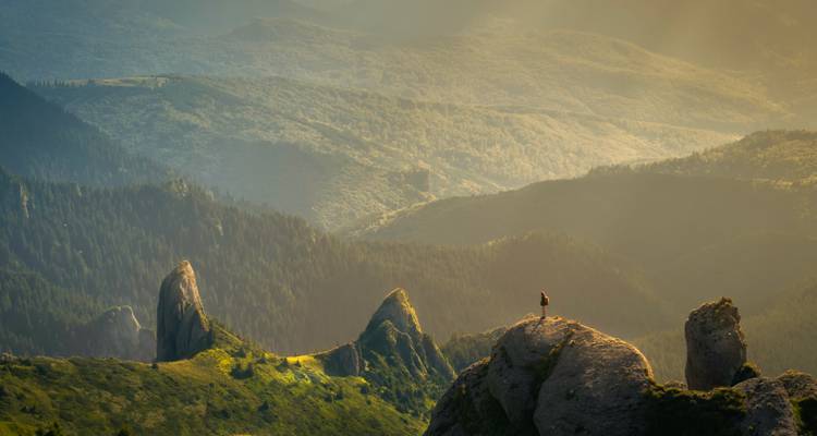 Lone hiker standing atop jagged rock spires overlooking vast misty mountain valleys lit by golden light.