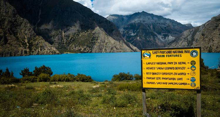 Een houten bord dat de kenmerken van Shey Phoksundo Nationaal Park belicht met een meer op de achtergrond.