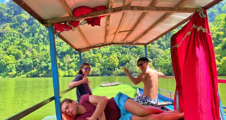 Twee mannen op een houten boot omringd door kalm groen water en weelderige groene heuvels.