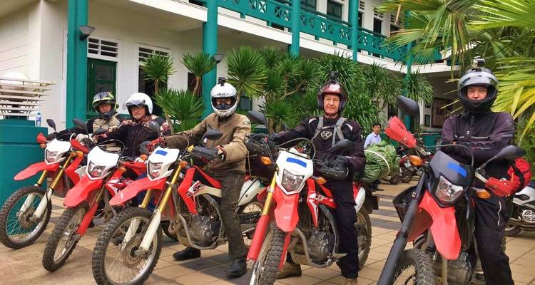 Gruppe von Motorradfahrern, die vor einem Gebäude mit üppigem Grün posieren.