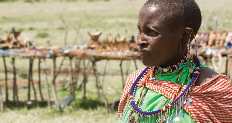 Maasai woman in vibrant traditional attire and beaded jewelry standing near a handcrafted market stall.