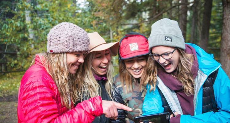 Quatre jeunes femmes en équipement de plein air riant ensemble en regardant un téléphone dans une clairière forestière.
