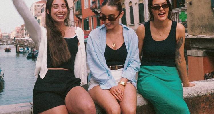 Three friends sit on canal wall laughing with colorful Venetian buildings behind