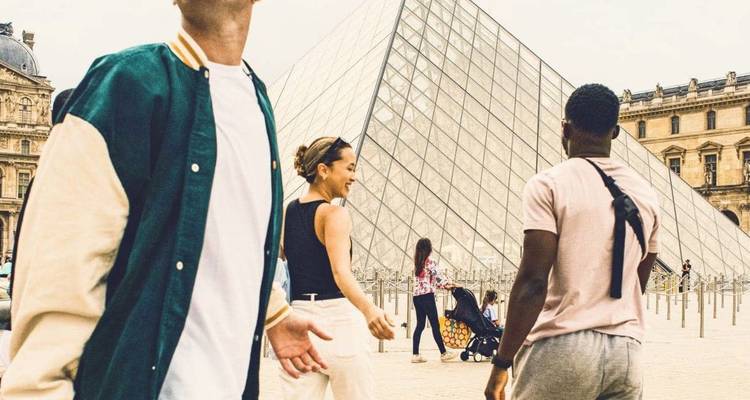 Group of friends laughing outside the Louvre Pyramid in Paris with glass structure and historic palace wings behind.