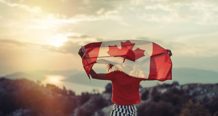 Un voyageur agite fièrement un grand drapeau canadien sur une crête de montagne au lever du soleil avec des lacs en contrebas.