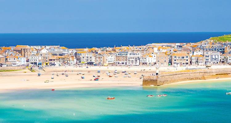Sunny aerial of St Ives harbour with turquoise waters, sandy beach, and rows of pastel cottages.